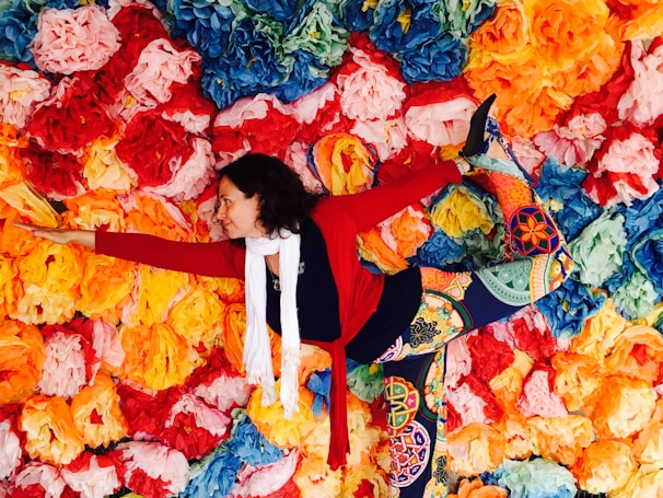 A person is performing a yoga pose, balancing against a vibrant backdrop of paper flowers in various colors including red, orange, yellow, blue, and green. They are wearing colorful, patterned leggings, a red sweater, and a white scarf.