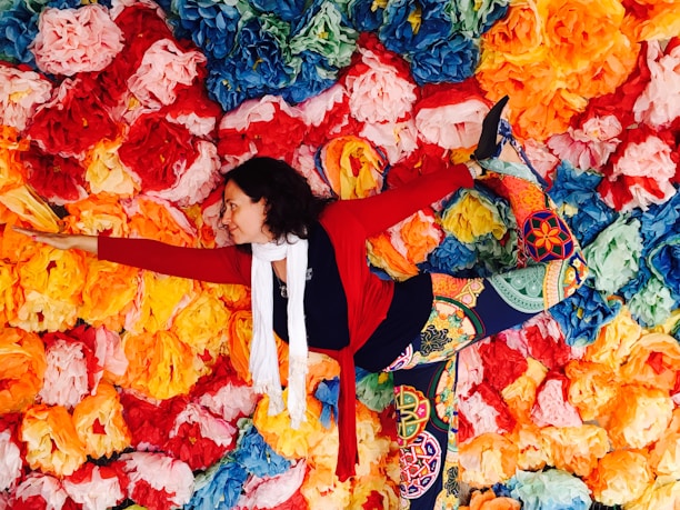 A person is performing a yoga pose, balancing against a vibrant backdrop of paper flowers in various colors including red, orange, yellow, blue, and green. They are wearing colorful, patterned leggings, a red sweater, and a white scarf.