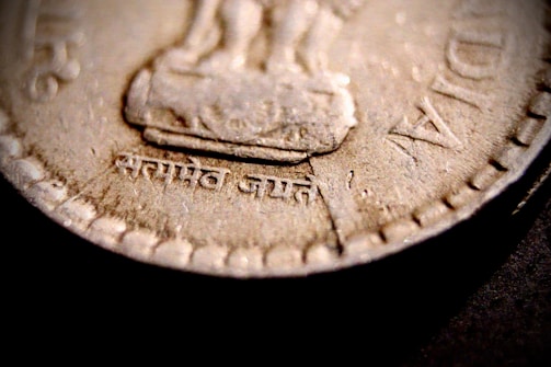 Close-up view of a coin featuring intricate patterns and inscriptions. The coin shows raised lettering in an unfamiliar script around its edges and a faint design in the middle.