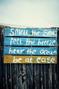A wooden sign with a rustic appearance features inspirational text painted in white. The sign has a background of blue and yellow planks, with the text highlighting sensory experiences associated with the sea.