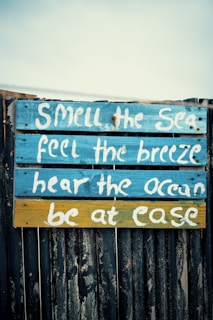 A wooden sign with a rustic appearance features inspirational text painted in white. The sign has a background of blue and yellow planks, with the text highlighting sensory experiences associated with the sea.
