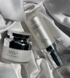 Two skincare product bottles are displayed on a textured, white fabric surface. One is a round jar labeled 'Multi Tea Serum', featuring a shiny silver lid. The other is a cylindrical bottle labeled 'Advanced Night Booster', also with silver accents.