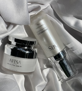 Two skincare product bottles are displayed on a textured, white fabric surface. One is a round jar labeled 'Multi Tea Serum', featuring a shiny silver lid. The other is a cylindrical bottle labeled 'Advanced Night Booster', also with silver accents.