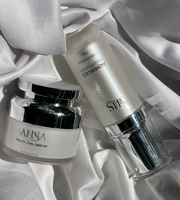Two skincare product bottles are displayed on a textured, white fabric surface. One is a round jar labeled 'Multi Tea Serum', featuring a shiny silver lid. The other is a cylindrical bottle labeled 'Advanced Night Booster', also with silver accents.