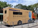 A mobile health services vehicle is parked on a street. The vehicle is beige with blue and orange accents, featuring text and logos, including 'Jockey Club Charles Kao Brain Health Services.' An illustrated portrait of a person in blue is displayed on the side. Trees and a building can be seen in the background, with metal barriers in front of the vehicle.