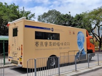 A mobile health services vehicle is parked on a street. The vehicle is beige with blue and orange accents, featuring text and logos, including 'Jockey Club Charles Kao Brain Health Services.' An illustrated portrait of a person in blue is displayed on the side. Trees and a building can be seen in the background, with metal barriers in front of the vehicle.