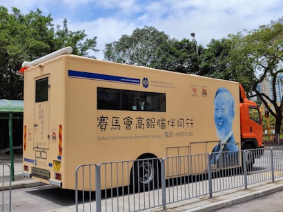 A mobile health services vehicle is parked on a street. The vehicle is beige with blue and orange accents, featuring text and logos, including 'Jockey Club Charles Kao Brain Health Services.' An illustrated portrait of a person in blue is displayed on the side. Trees and a building can be seen in the background, with metal barriers in front of the vehicle.