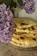 A stack of flatbread or pastry is placed on a newspaper, with pieces cut and stacked on top, revealing a filling of cheese and herbs. In the foreground, clusters of purple lilac flowers are visible, slightly out of focus, adding a touch of color and creating a rustic aesthetic.