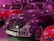 Several vintage cars are showcased in an indoor setting. The primary focus is on a grey classic car with a white and black license plate reading 'BAUJ 1963'. The car has a highly reflective surface, and the surroundings are lit with a purple hue. Behind and beside the grey car, other vintage cars are visible, including a red one.