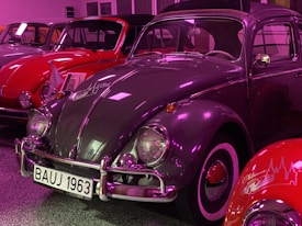 Several vintage cars are showcased in an indoor setting. The primary focus is on a grey classic car with a white and black license plate reading 'BAUJ 1963'. The car has a highly reflective surface, and the surroundings are lit with a purple hue. Behind and beside the grey car, other vintage cars are visible, including a red one.