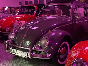 Several vintage cars are showcased in an indoor setting. The primary focus is on a grey classic car with a white and black license plate reading 'BAUJ 1963'. The car has a highly reflective surface, and the surroundings are lit with a purple hue. Behind and beside the grey car, other vintage cars are visible, including a red one.