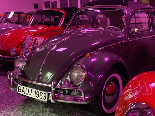 Several vintage cars are showcased in an indoor setting. The primary focus is on a grey classic car with a white and black license plate reading 'BAUJ 1963'. The car has a highly reflective surface, and the surroundings are lit with a purple hue. Behind and beside the grey car, other vintage cars are visible, including a red one.