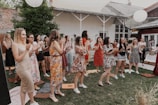 A group of people, mostly women, are gathered outdoors in a garden or yard setting. They are standing on grass, some clapping and appearing to engage in a celebratory or cheerful activity. The attendees are dressed in a variety of colorful summer outfits. The setting includes elements like a brick wall, wooden structures, and paper lanterns hanging above.