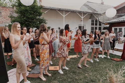 A group of people, mostly women, are gathered outdoors in a garden or yard setting. They are standing on grass, some clapping and appearing to engage in a celebratory or cheerful activity. The attendees are dressed in a variety of colorful summer outfits. The setting includes elements like a brick wall, wooden structures, and paper lanterns hanging above.