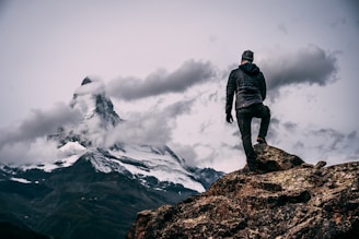 A person stands on a rocky outcrop, gazing at a snow-capped mountain peak partially shrouded by clouds. The scene conveys a sense of adventure and awe.