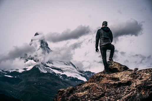 A person stands on a rocky outcrop, gazing at a snow-capped mountain peak partially shrouded by clouds. The scene conveys a sense of adventure and awe.