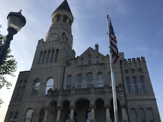 A historic stone building featuring a prominent clock tower rises under a clear blue sky. Its architectural design includes arched windows and a mixture of rectangular and circular shapes. In the foreground, an American flag is raised on a pole, gently waving, while a vintage streetlamp stands to the side, framed by leafy green tree branches.