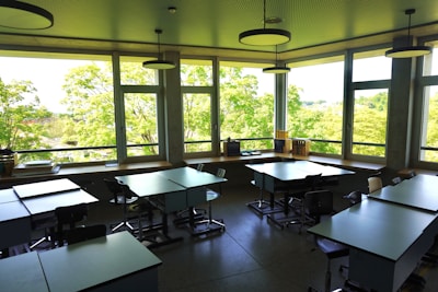 A spacious classroom is filled with several tables and chairs, illuminated by natural light streaming through large windows. Outside, lush green trees create a vibrant backdrop. The room is neatly arranged, with no students present, and has books and supplies on the windowsills.