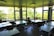 A spacious classroom is filled with several tables and chairs, illuminated by natural light streaming through large windows. Outside, lush green trees create a vibrant backdrop. The room is neatly arranged, with no students present, and has books and supplies on the windowsills.