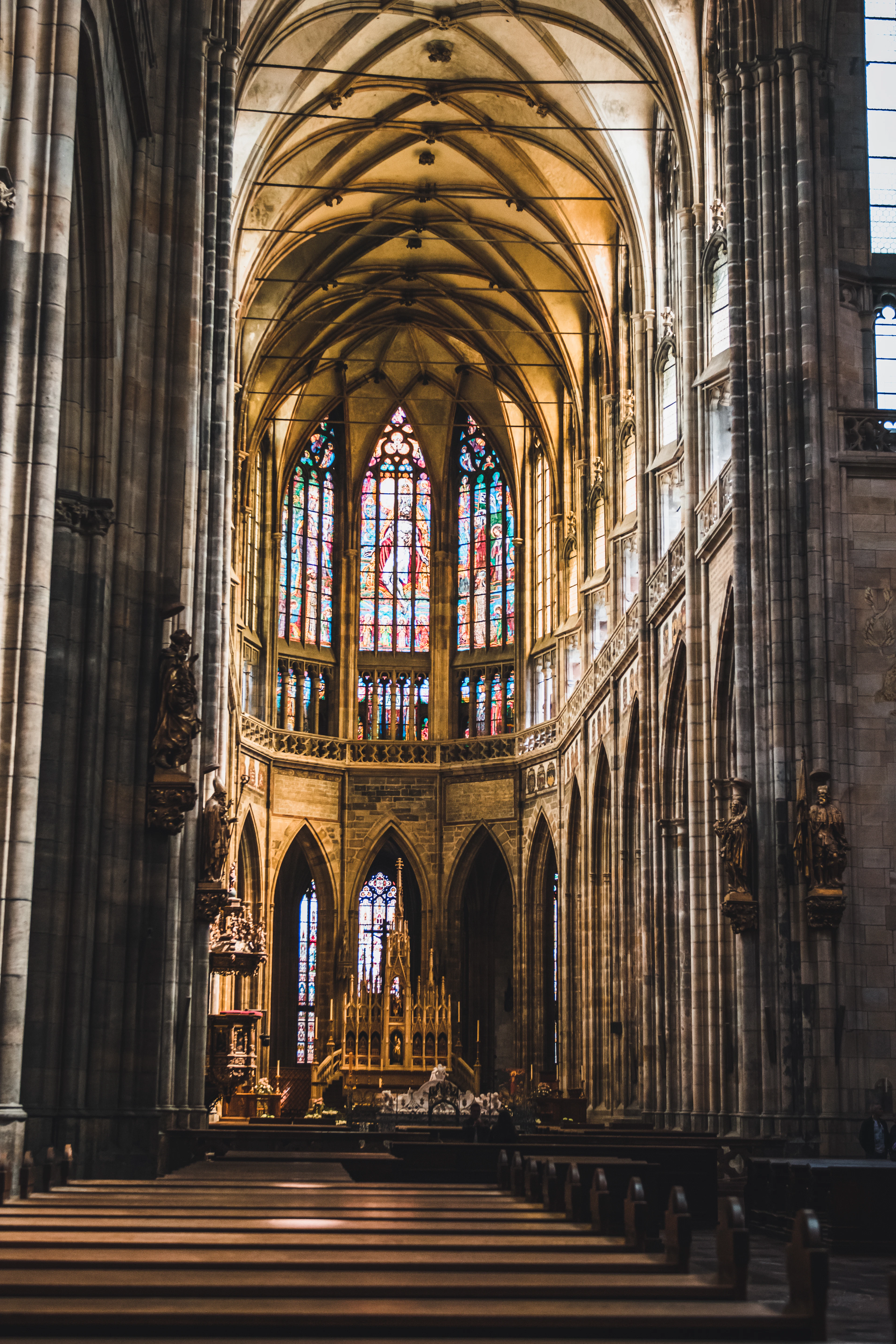 The nave of St Vitus Cathedral in Prague, ribbed stone vaults soaring above empty wooden pews towards a wall of jewel-coloured stained glass and the golden high altar.
