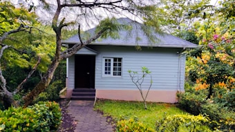 A small, light-blue house with a pitched roof is surrounded by lush greenery. The yard features various plants and a path leading to the front door. Trees and shrubs frame the house, creating a secluded and peaceful atmosphere.