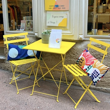 Two yellow metal chairs with colorful cushions are placed around a small yellow metal table. A potted succulent sits on the table along with some papers. This setup is in front of a shop with a stone wall, and there are multiple signs in the window promoting a vintage clothing store located upstairs.