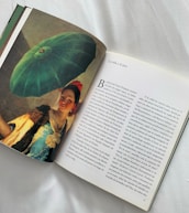 An open book lies on a light-colored surface, featuring a page with a painting on the left and text on the right. The painting shows a woman in traditional attire holding a large green umbrella. She has a warm expression and is dressed in colorful clothing, with a red accessory in her hair. The right page contains printed text in Spanish, discussing art and life.