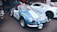 A blue vintage racing car with the number 110 on the side is parked on the street. It features branding stickers such as 'Cibie' and has distinctive headlamps and a rally badge on the hood. Behind it, a modern white sports car is partially visible among a group of people, suggesting a car show or gathering.