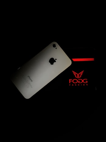 An iPhone is placed against a dark background, partially overlapping packaging with red and white branding that reads 'FOGG FASHION'.