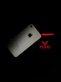An iPhone is placed against a dark background, partially overlapping packaging with red and white branding that reads 'FOGG FASHION'.