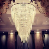 An opulent chandelier composed of multiple cascading layers of crystal strands, suspended from a large circular frame. The chandelier is intricately detailed and emits a soft glow that illuminates the ornate ceiling designs. The surrounding area includes elegant drapery and classic architectural elements.