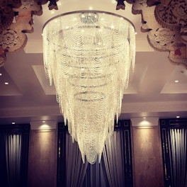 An opulent chandelier composed of multiple cascading layers of crystal strands, suspended from a large circular frame. The chandelier is intricately detailed and emits a soft glow that illuminates the ornate ceiling designs. The surrounding area includes elegant drapery and classic architectural elements.
