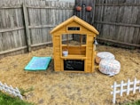 A small wooden playhouse resembling a café is situated in a backyard. The structure has a chalkboard sign with a menu, a bell hanging on the side, and a small bucket placed on the counter. Two soft, patterned beanbag chairs are nearby, along with a light blue mat with decorative patterns. The area is fenced with wooden planks and there are small white decorative fences on the ground. Two wall planters hang on the fence, adding a touch of greenery.