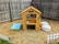 A small wooden playhouse resembling a café is situated in a backyard. The structure has a chalkboard sign with a menu, a bell hanging on the side, and a small bucket placed on the counter. Two soft, patterned beanbag chairs are nearby, along with a light blue mat with decorative patterns. The area is fenced with wooden planks and there are small white decorative fences on the ground. Two wall planters hang on the fence, adding a touch of greenery.