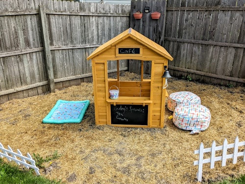A small wooden playhouse resembling a caf&eacute; is situated in a backyard. The structure has a chalkboard sign with a menu, a bell hanging on the side, and a small bucket placed on the counter. Two soft, patterned beanbag chairs are nearby, along with a light blue mat with decorative patterns. The area is fenced with wooden planks and there are small white decorative fences on the ground. Two wall planters hang on the fence, adding a touch of greenery.