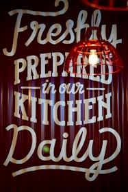 A sign with bold, white text on a dark background featuring the message 'Freshly Prepared in our Kitchen Daily.' A red industrial-style pendant light with an exposed bulb hangs in the foreground.