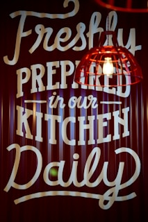A sign with bold, white text on a dark background featuring the message 'Freshly Prepared in our Kitchen Daily.' A red industrial-style pendant light with an exposed bulb hangs in the foreground.