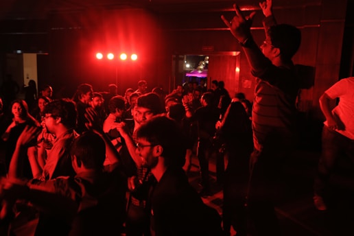 A lively group of people dancing in a dimly lit room with red lighting. Many are engaged in animated conversation and movement, with one person on the right raising their hands enthusiastically. The atmosphere suggests a festive or party setting.