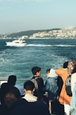 A group of people is gathered on the deck of a boat, looking out at the sea. A yacht cruises by in the background, and a cityscape with densely packed buildings is visible across the water. The atmosphere is calm and leisurely, with clear skies and gentle waves.