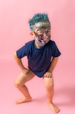 A young child stands confidently with hands on their knees, hair styled with blue dye, and face playfully covered in colorful paint. The background is a solid pink, enhancing the vibrant and playful atmosphere.