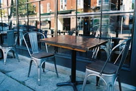 A rustic wooden table is flanked by metal chairs on a sidewalk in front of a large window, reflecting the urban street scene and neighboring buildings. The setup appears to belong to an outdoor seating area of a cafe or restaurant.
