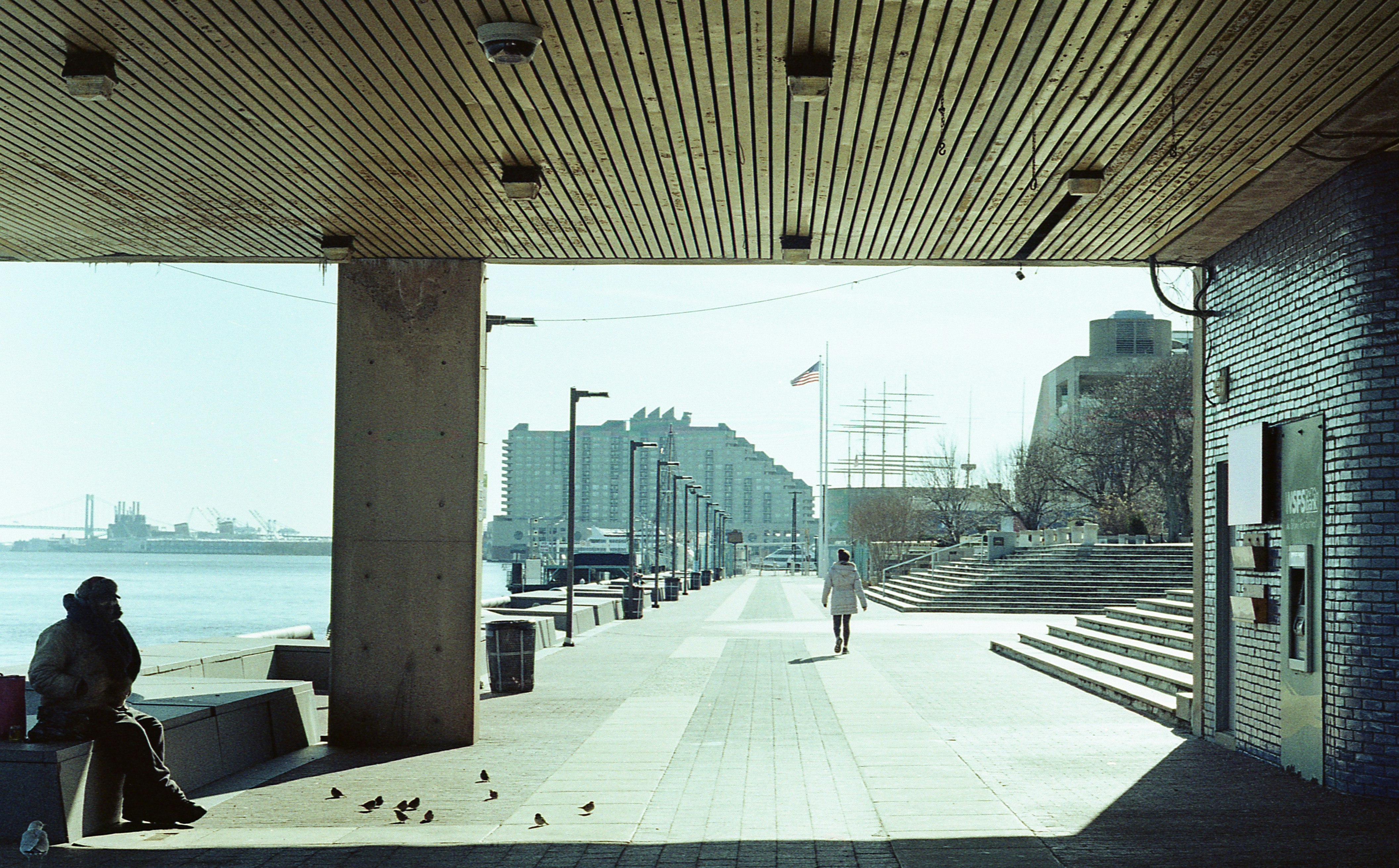 a person sitting on a bench next to a body of water, Philly pier. Shot on Minolta SRT-101.
