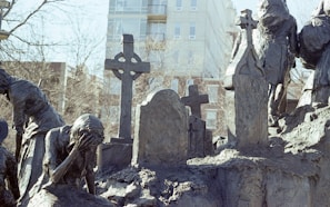 A somber sculpture scene depicting figures in mourning. The figures are made of a dark, rough-textured material and appear to be gathered around a graveyard setting, with one figure holding their head in their hand in a gesture of grief. Several gravestones and crosses are visible, adding to the somber atmosphere.