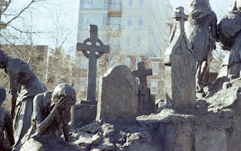 A somber sculpture scene depicting figures in mourning. The figures are made of a dark, rough-textured material and appear to be gathered around a graveyard setting, with one figure holding their head in their hand in a gesture of grief. Several gravestones and crosses are visible, adding to the somber atmosphere.