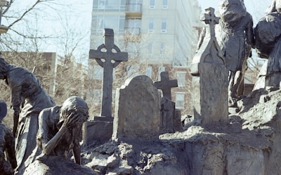 A somber sculpture scene depicting figures in mourning. The figures are made of a dark, rough-textured material and appear to be gathered around a graveyard setting, with one figure holding their head in their hand in a gesture of grief. Several gravestones and crosses are visible, adding to the somber atmosphere.