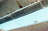 A metal gutter is attached to the edge of a corrugated roof, with water droplets dripping down. The background shows a blurred brick wall and a blue sky.