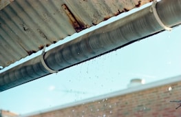 A metal gutter is attached to the edge of a corrugated roof, with water droplets dripping down. The background shows a blurred brick wall and a blue sky.