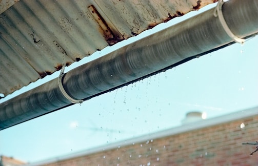 A metal gutter is attached to the edge of a corrugated roof, with water droplets dripping down. The background shows a blurred brick wall and a blue sky.