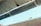 A metal gutter is attached to the edge of a corrugated roof, with water droplets dripping down. The background shows a blurred brick wall and a blue sky.