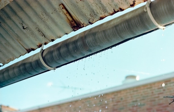 A metal gutter is attached to the edge of a corrugated roof, with water droplets dripping down. The background shows a blurred brick wall and a blue sky.